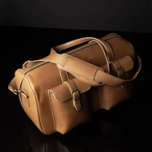 Angled view of Marcella Overnight Duffle Bag in tan leather, showcasing reinforced carry handles and brass hardware.